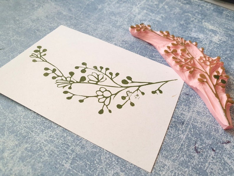 May include: A green floral design stamp with a pink rubber base. The stamp is used to create a floral design on paper.