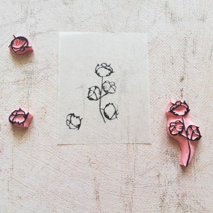 May include: A collection of floral stamps and a stamped image on paper. The stamps are pink with black floral designs. The stamped image shows a stem with several flowers. The background is a textured, off-white surface.