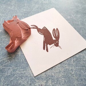 May include: Two brown wooden stamps of rabbits. One stamp shows a rabbit standing on its hind legs with its front paws raised. The other stamp shows two rabbits stacked on top of each other.