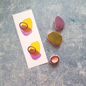 May include: Four rubber stamps with abstract shapes. Two stamps are yellow, pink, and brown. One stamp is green. One stamp is purple.