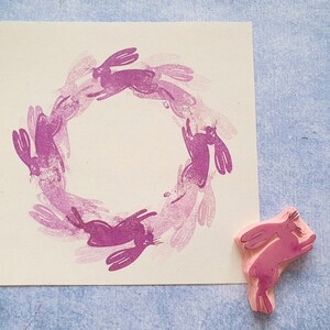 May include: A pink rubber stamp with a design of a circle of bunnies. The stamp is on a white background.