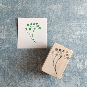 May include: A wooden stamp with a green floral design. The stamp is on a light blue surface. The design is of a small branch with leaves.