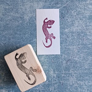 May include: A wooden stamp with a lizard design next to a print of the same lizard in purple ink on white paper. The stamp is rectangular and the lizard is in a side profile. The background is a textured blue surface.