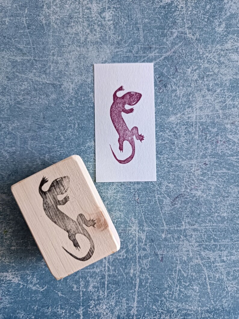 May include: A wooden stamp with a lizard design next to a print of the same lizard in purple ink on white paper. The stamp is rectangular and the lizard is in a side profile. The background is a textured blue surface.