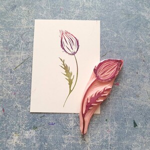 May include: A pink rubber stamp with a detailed design of a flower bud and stem. The stamp is used to create a print of a flower on paper.