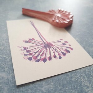May include: A purple and pink dandelion flower stamp with a wooden handle. The stamp is used to create a decorative design on paper.