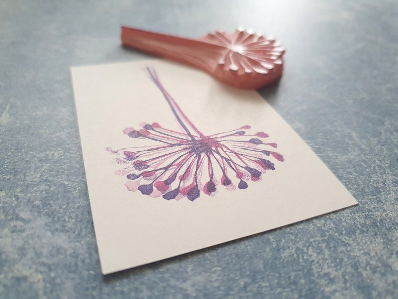 May include: A purple and pink dandelion flower stamp with a wooden handle. The stamp is used to create a decorative design on paper.