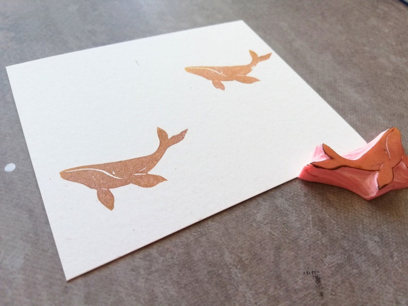 May include: Two brown whale stamps on a white card with a pink rubber whale stamp on a gray surface.
