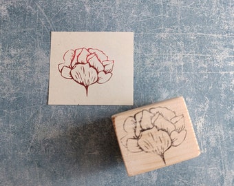 Könnte beinhalten: Ein Holzstempel mit einem roten floralen Design einer einzelnen Blume mit mehreren Blütenblättern. Der Stempel befindet sich auf einer blauen, strukturierten Oberfläche.