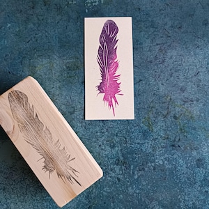 Può includere: Un timbro in legno con un design inciso di una piuma. Il timbro è mostrato accanto a un'immagine stampata di una piuma in tonalità di rosa e viola.