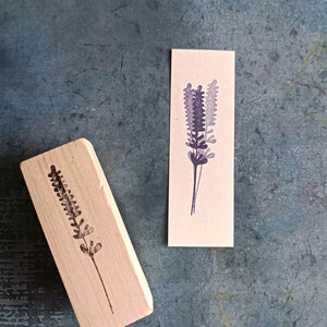 May include: Two wooden stamps with a lavender design. The stamps are on a textured blue surface. The design is a simple silhouette of a lavender flower.