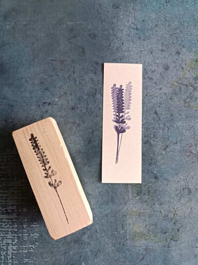 May include: Two wooden stamps with a lavender design. The stamps are on a textured blue surface. The design is a simple silhouette of a lavender flower.
