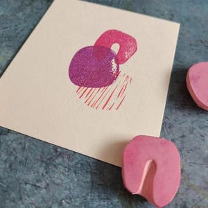 May include: A cream-colored card with a magenta abstract print of two overlapping shapes and red lines. Pink rubber stamps are scattered around the card on a textured blue surface. The stamps have different shapes.