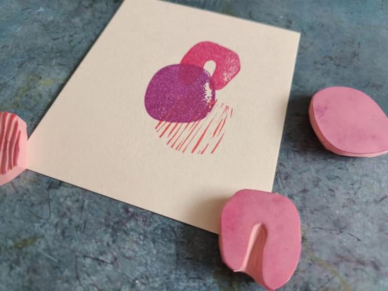 May include: A cream-colored card with a magenta abstract print of two overlapping shapes and red lines. Pink rubber stamps are scattered around the card on a textured blue surface. The stamps have different shapes.