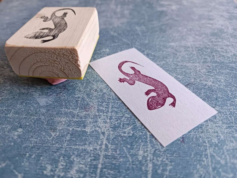 May include: A wooden stamp with a lizard design and a matching stamped image on a white rectangle. The stamp is rectangular with a light wood grain. The lizard image is dark and detailed.