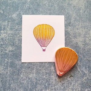 Pode incluir: Um carimbo de madeira de um bal&atilde;o de ar quente com um degrad&ecirc; amarelo e rosa. O carimbo est&aacute; em um cart&atilde;o branco com uma imagem impressa de um bal&atilde;o de ar quente.
