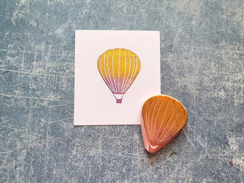 Pode incluir: Um carimbo de madeira de um bal&atilde;o de ar quente com um degrad&ecirc; amarelo e rosa. O carimbo est&aacute; em um cart&atilde;o branco com uma imagem impressa de um bal&atilde;o de ar quente.
