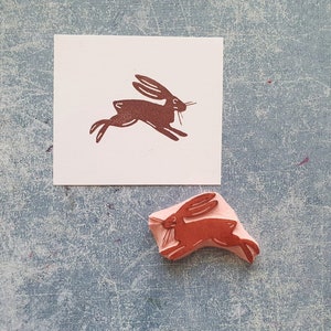 May include: A brown rubber stamp with a design of a running rabbit. The stamp is on a white card with a brown imprint of the rabbit design.
