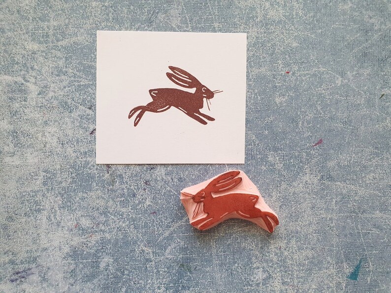 May include: A brown rubber stamp with a design of a running rabbit. The stamp is on a white card with a brown imprint of the rabbit design.