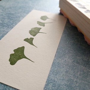 May include: A white card with a row of five green ginkgo leaf prints. The leaves are arranged in a line, getting progressively larger from left to right. The card is on a blue textured surface.