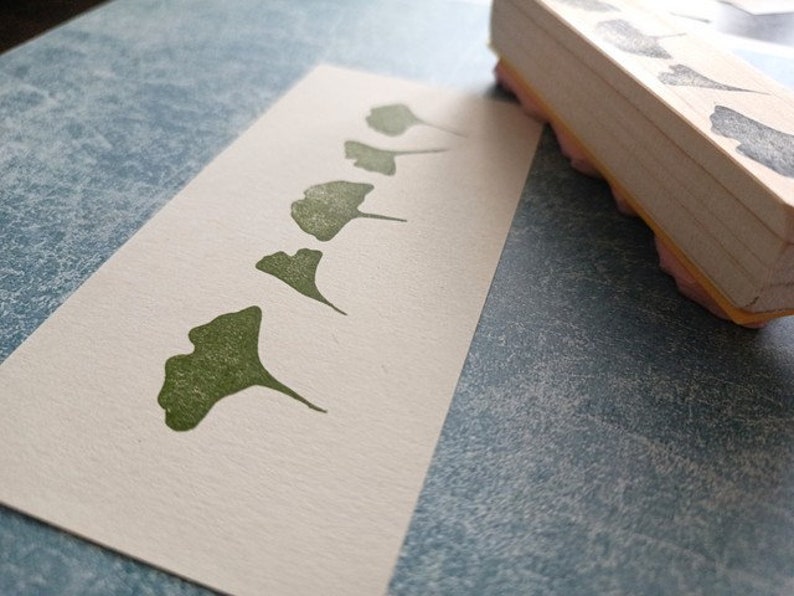 May include: A white card with a row of five green ginkgo leaf prints. The leaves are arranged in a line, getting progressively larger from left to right. The card is on a blue textured surface.