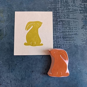 Può includere: Un timbro di gomma in legno con un contorno giallo di un coniglio seduto su un cartoncino bianco. Il timbro è marrone e presenta un design dettagliato.