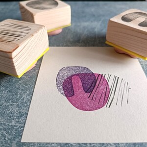 May include: Wooden art stamps with geometric designs, including lines, circles, and abstract shapes. The stamps are on pink pads with yellow bases. A stamped image in shades of purple and black is on a white paper.