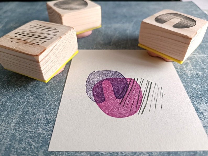 May include: Wooden art stamps with geometric designs, including lines, circles, and abstract shapes. The stamps are on pink pads with yellow bases. A stamped image in shades of purple and black is on a white paper.