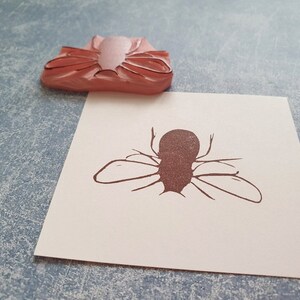 May include: A pink rubber stamp of a fly rests above a paper with a brown fly illustration. The stamp is a light pink color, and the fly illustration is brown. The paper is white.