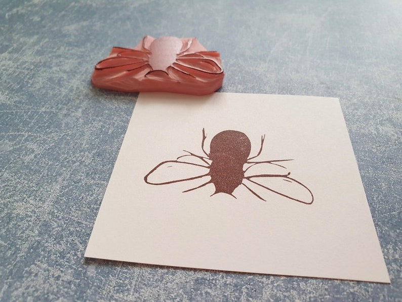 May include: A pink rubber stamp of a fly rests above a paper with a brown fly illustration. The stamp is a light pink color, and the fly illustration is brown. The paper is white.