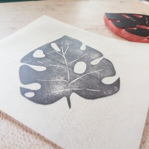 May include: A gray leaf print on white paper, created with a stamp. The stamp, with a black leaf design, is visible in the background. The leaf print has a detailed vein structure and several holes.