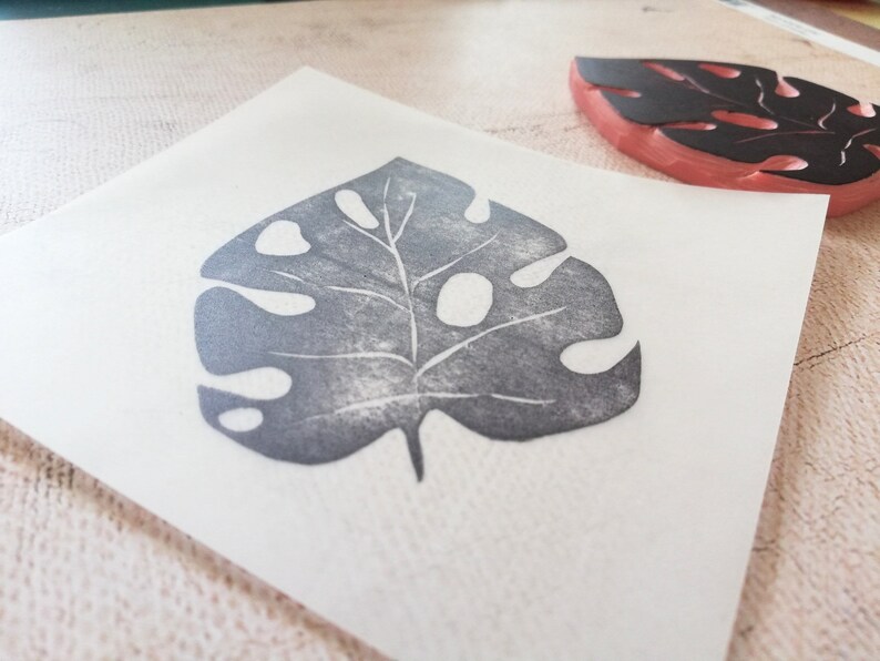 May include: A gray leaf print on white paper, created with a stamp. The stamp, with a black leaf design, is visible in the background. The leaf print has a detailed vein structure and several holes.