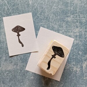 K&ouml;nnte beinhalten: Ein Stempel aus Holz mit einer schwarzen Silhouette eines Pilzes. Der Stempel befindet sich auf einer wei&szlig;en Oberfl&auml;che.