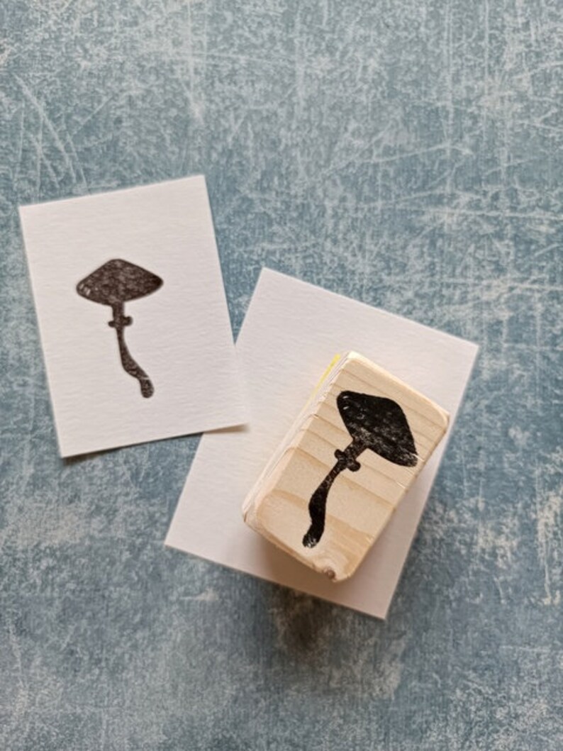 K&ouml;nnte beinhalten: Ein Stempel aus Holz mit einer schwarzen Silhouette eines Pilzes. Der Stempel befindet sich auf einer wei&szlig;en Oberfl&auml;che.