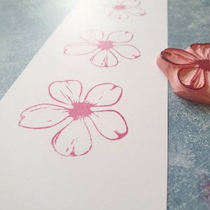 Pode incluir: Um close-up de um carimbo de borracha rosa com um desenho de flor. O carimbo est&aacute; em uma superf&iacute;cie branca com v&aacute;rias impress&otilde;es de flores rosa.