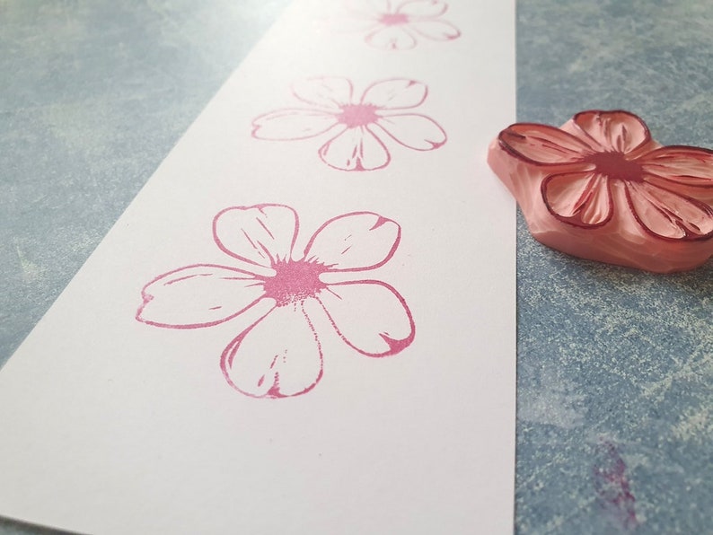Pode incluir: Um close-up de um carimbo de borracha rosa com um desenho de flor. O carimbo est&aacute; em uma superf&iacute;cie branca com v&aacute;rias impress&otilde;es de flores rosa.