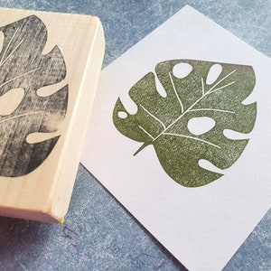 May include: A wooden stamp with a leaf design and a green leaf print on white paper. The stamp is made of light-colored wood and features a detailed carving of a monstera leaf. The printed leaf is a shade of green with visible texture.