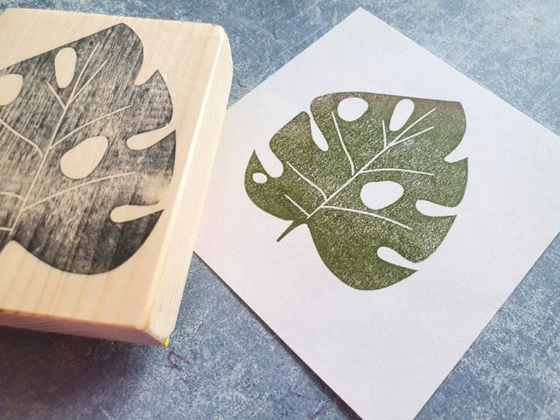 May include: A wooden stamp with a leaf design and a green leaf print on white paper. The stamp is made of light-colored wood and features a detailed carving of a monstera leaf. The printed leaf is a shade of green with visible texture.