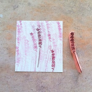 May include: A wooden stamp with a design of a lavender flower. The stamp is used to create a pattern of lavender flowers on a piece of paper.