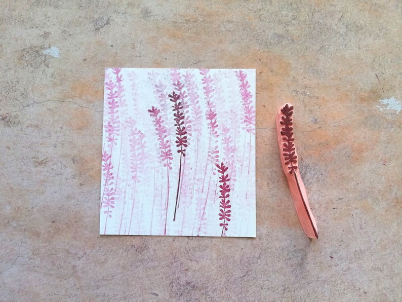 May include: A wooden stamp with a design of a lavender flower. The stamp is used to create a pattern of lavender flowers on a piece of paper.