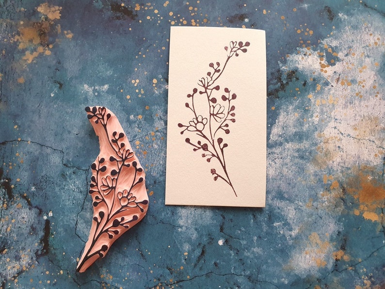 May include: A wooden stamp with a floral design, and a card with a floral design printed on it. The stamp is pink and the card is cream.