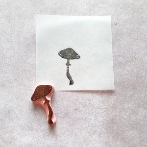 K&ouml;nnte beinhalten: Ein brauner Gummistempel in Form eines Pilzes. Der Stempel wurde verwendet, um einen Abdruck eines Pilzes auf einem wei&szlig;en Blatt Papier zu erstellen.