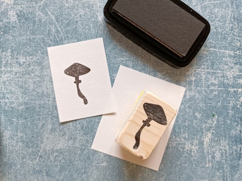 K&ouml;nnte beinhalten: Zwei Holzstempel mit einem Pilzmotiv. Die Stempel liegen auf wei&szlig;em Papier und ein schwarzes Stempelkissen befindet sich im Hintergrund.