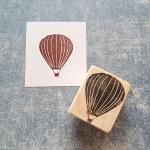Pode incluir: Um carimbo de madeira com uma silhueta preta de um bal&atilde;o de ar quente. O bal&atilde;o tem uma cesta e &eacute; delineado com linhas brancas.