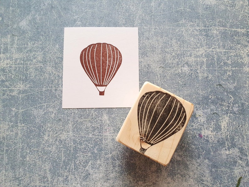 Pode incluir: Um carimbo de madeira com uma silhueta preta de um bal&atilde;o de ar quente. O bal&atilde;o tem uma cesta e &eacute; delineado com linhas brancas.