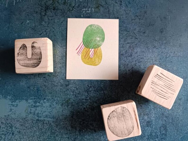 May include: An overhead shot shows a collection of wooden stamps and a small print on a textured blue surface. The print features green and yellow abstract shapes with red lines. The stamps have various black designs.