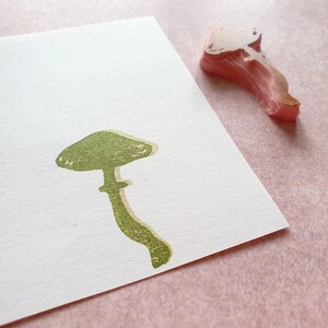 K&ouml;nnte beinhalten: Ein gr&uuml;ner Gummistempel mit einem Pilz mit abgerundetem Hut und Stiel. Der Stempel befindet sich auf einem wei&szlig;en Blatt Papier.