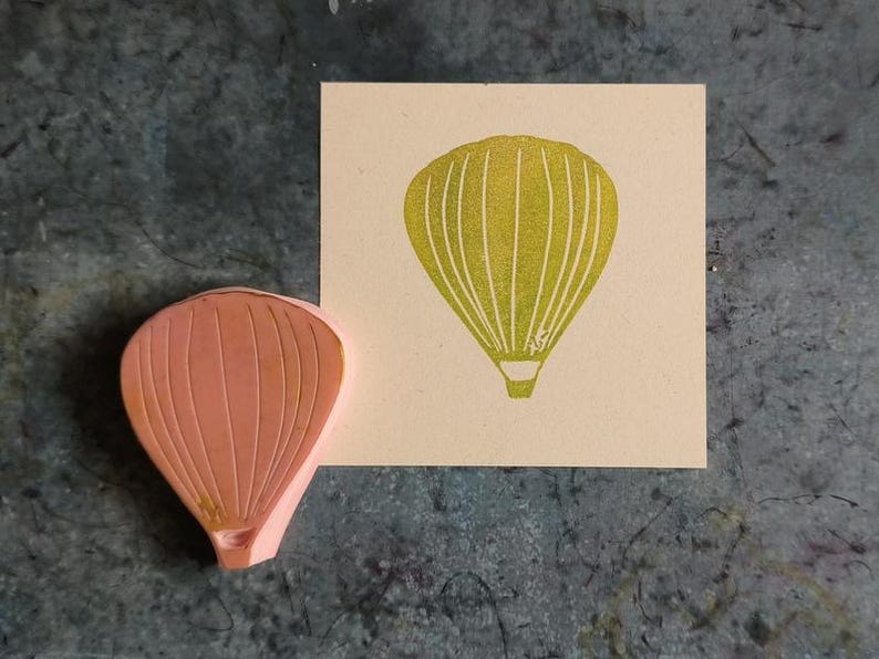Pode incluir: Um carimbo de borracha rosa com um desenho de bal&atilde;o de ar quente. O carimbo &eacute; usado para criar uma imagem de bal&atilde;o de ar quente amarelo.