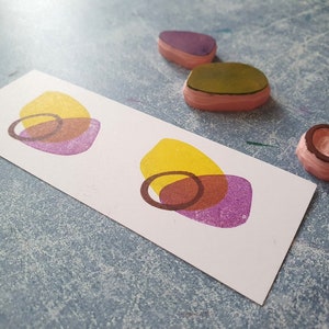 May include: Two sets of overlapping circles in yellow, brown, and purple on a white card. The circles are printed with a rubber stamp.