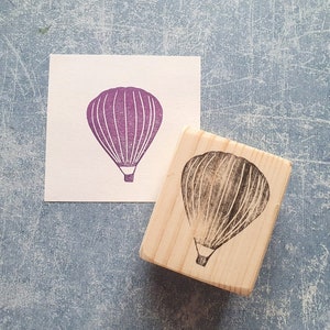 Pode incluir: Um carimbo de bloco de madeira com um desenho de bal&atilde;o de ar quente roxo. O carimbo est&aacute; em um cart&atilde;o branco com um desenho de bal&atilde;o de ar quente roxo.